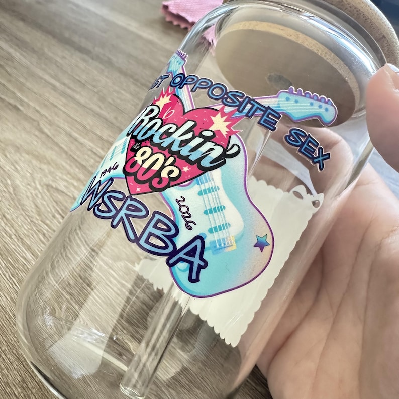 May include: A clear glass tumbler with a bamboo lid and glass straw, featuring a colorful decal of a light blue electric guitar, a pink heart, and the text "Rockin' 80's", "WSRBA", "1946", "2026", and "OPPOSITE SEX" in bold, stylized lettering.