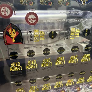May include: A clear sheet of adhesive labels featuring the text "LEMON DROP" in yellow, "BARRSIPS" in black, and "BUCKZ BARR" in small circles. The sheet includes red circular "ROJA de Tere" logos and a shield-shaped emblem with a gold helmet.