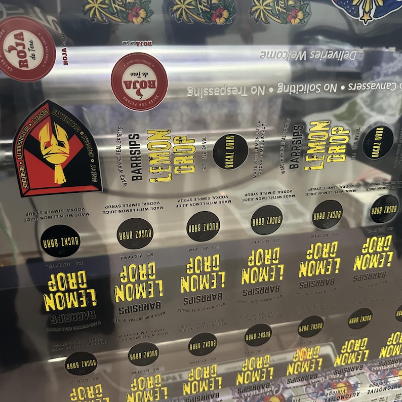 May include: A clear sheet of adhesive labels featuring the text "LEMON DROP" in yellow, "BARRSIPS" in black, and "BUCKZ BARR" in small circles. The sheet includes red circular "ROJA de Tere" logos and a shield-shaped emblem with a gold helmet.