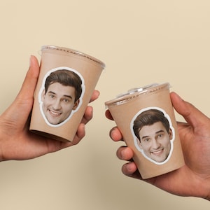 May include: Two takeaway coffee cups with clear lids, each featuring a sticker of a smiling person's face. The cups are made of brown paper and are held in hands against a neutral background.