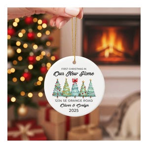 May include: A white ceramic Christmas ornament with a gold string, featuring text that reads "First Christmas in Our New Home." Below are illustrated Christmas trees and the address "1234 SE Orange Road, Oliver & Evelyn, 2025."