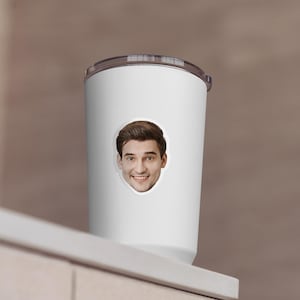 May include: White travel mug with a smiling face sticker. The mug has a silver lid and a white body. The sticker features a man with brown hair and a friendly expression. The mug is on a white surface.