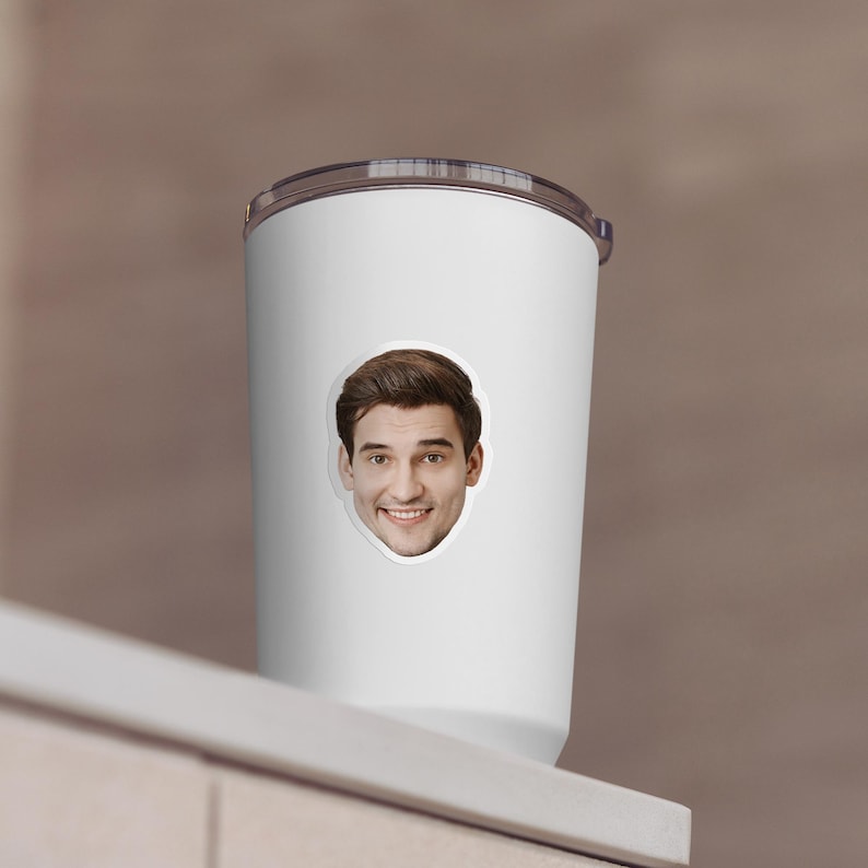 May include: White travel mug with a smiling face sticker. The mug has a silver lid and a white body. The sticker features a man with brown hair and a friendly expression. The mug is on a white surface.