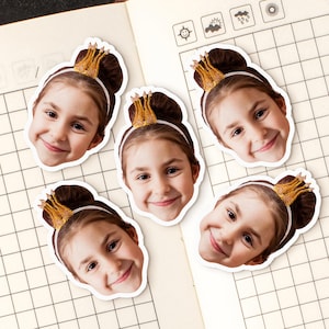 May include: Five stickers of a smiling girl with brown hair in a bun, wearing a gold crown and a white headband. The stickers are on a notebook with a grid pattern.