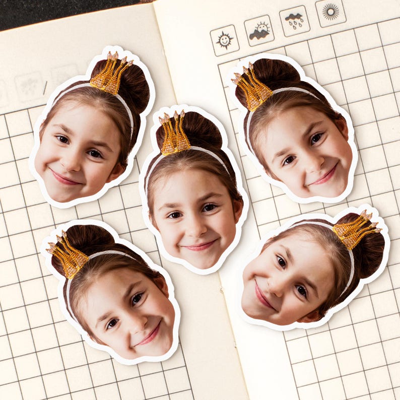 May include: Five stickers of a smiling girl with brown hair in a bun, wearing a gold crown and a white headband. The stickers are on a notebook with a grid pattern.