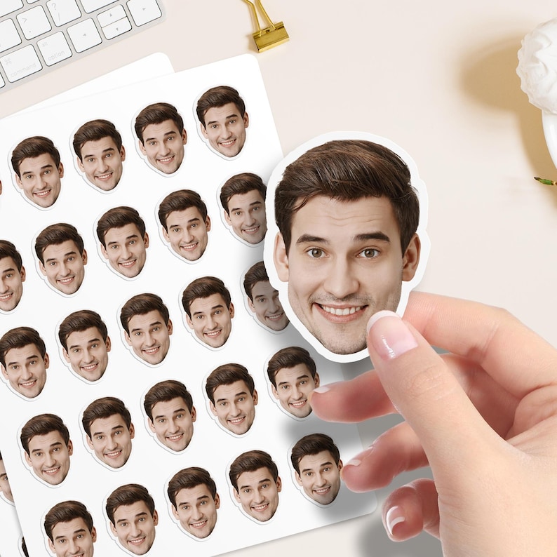 May include: A sheet of stickers with a smiling man's face. The stickers are white with a full-colour image of the man's face. One sticker is being held by a hand. A keyboard and office supplies are in the background.
