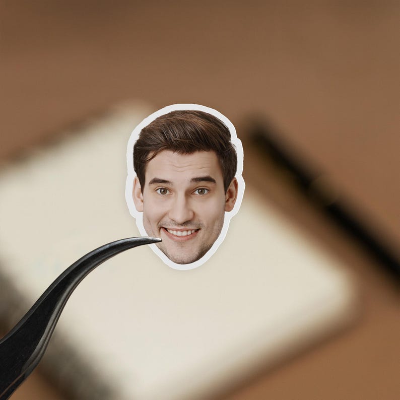 May include: A sticker of a smiling man's face, with brown hair and a white border. The sticker is held by black tweezers, against a blurred background of a notebook and pen.