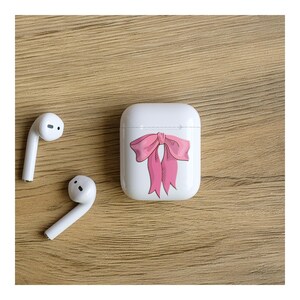 May include: White wireless earbuds and a white charging case with a pink bow design. The earbuds and case are on a wooden surface. The bow is a hand-drawn style with pink and red accents.