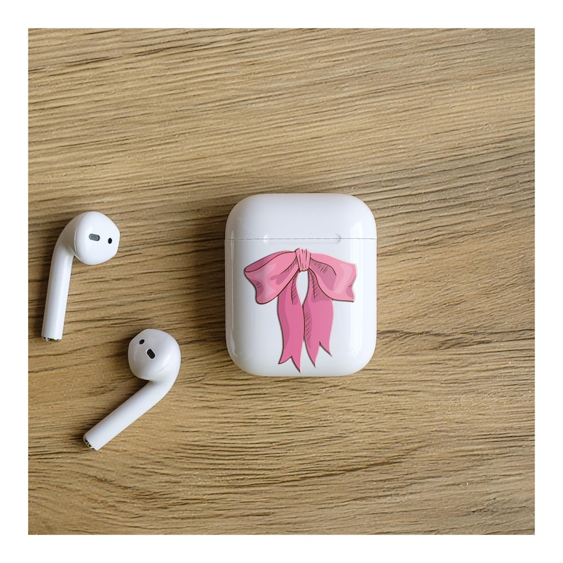 May include: White wireless earbuds and a white charging case with a pink bow design. The earbuds and case are on a wooden surface. The bow is a hand-drawn style with pink and red accents.