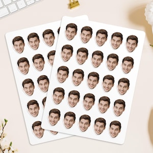 May include: Two sheets of white stickers featuring a smiling man's face. Each sheet contains multiple stickers with a close-up of the man's face. The stickers are oval-shaped and have a glossy finish. The background is a light beige colour.