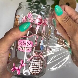 May include: A clear glass can-shaped tumbler featuring a colorful wrap-around design with illustrations of a teddy bear, pink tulips, a pink bow, a pink quilted handbag, a lipstick tube, a pink balloon dog, and a disco ball.