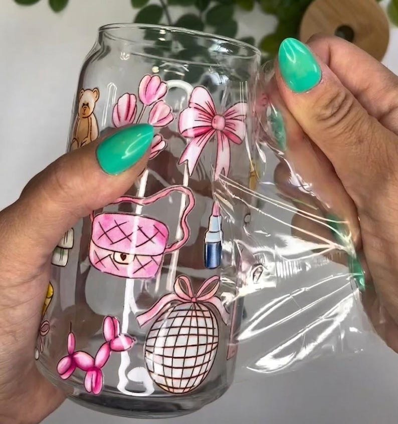 May include: A clear glass can-shaped tumbler featuring a colorful wrap-around design with illustrations of a teddy bear, pink tulips, a pink bow, a pink quilted handbag, a lipstick tube, a pink balloon dog, and a disco ball.