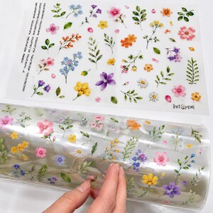 May include: A clear adhesive sheet and a matching roll of transparent film, both featuring a repeating pattern of watercolor-style illustrations of various small flowers, buds, and green leaves in shades of pink, yellow, blue, and purple.