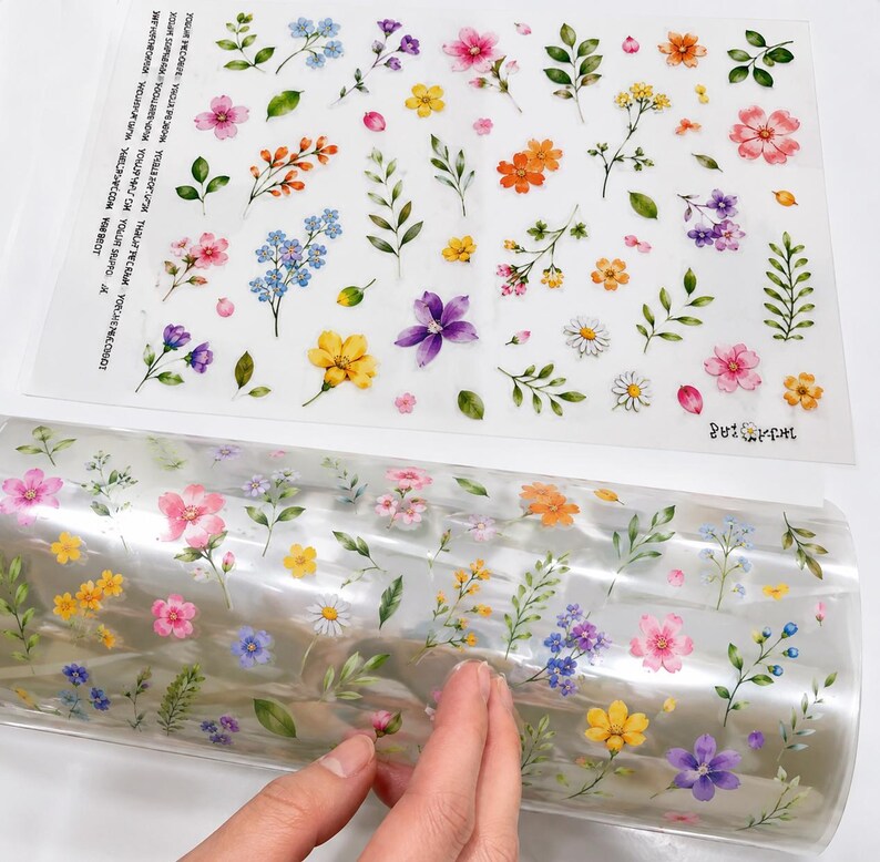 May include: A clear adhesive sheet and a matching roll of transparent film, both featuring a repeating pattern of watercolor-style illustrations of various small flowers, buds, and green leaves in shades of pink, yellow, blue, and purple.