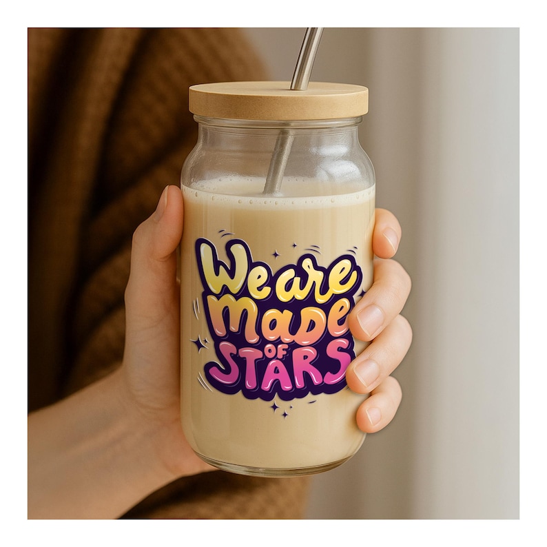 May include: A clear glass jar with a wooden lid and a metal straw, filled with a creamy beverage. The jar features the text "We are made of Stars" in colorful bubble letters. The jar is held by a person.