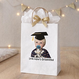 May include: White gift bag with a photo of a baby wearing a graduation cap and holding a stuffed animal. The bag has a gold bow and the text "Look Who's Graduated". The bag is filled with white tissue paper.