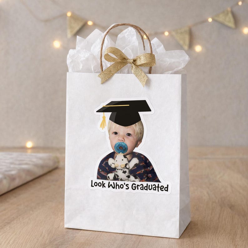 May include: White gift bag with a photo of a baby wearing a graduation cap and holding a stuffed animal. The bag has a gold bow and the text "Look Who's Graduated". The bag is filled with white tissue paper.