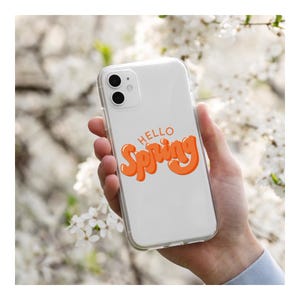 May include: A clear phone case with the words "HELLO Spring" in orange script. The phone case is held in a person's hand, with a blurred background of white flowers.