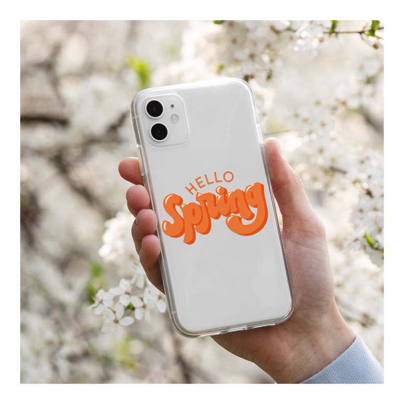 May include: A clear phone case with the words "HELLO Spring" in orange script. The phone case is held in a person's hand, with a blurred background of white flowers.