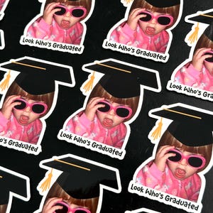May include: Sticker sheet featuring multiple stickers of a child wearing pink sunglasses and a graduation cap. The stickers have the text "Look Who's Graduated" in white.