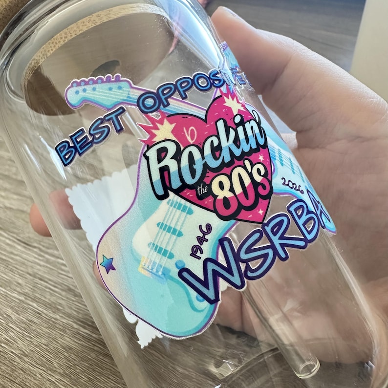 May include: A clear glass tumbler with a bamboo lid and a glass straw, featuring a colorful decal of a light blue electric guitar, a pink heart, and the text "BEST OPPOSITES", "Rockin the 80's", "1946", "2026", and "WSRBA" in bold lettering.