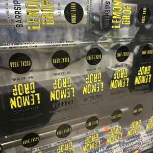 May include: A sheet of clear adhesive labels for Barrsips Lemon Drop vodka cocktails. Each label features the yellow text Lemon Drop, the brand name Buckz Barr in a black circle, and the text Made with lemon juice, vodka, simple syrup.