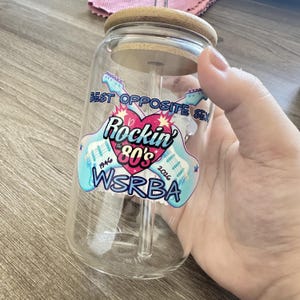 May include: A clear glass tumbler with a bamboo lid and a glass straw. The front features a graphic with two light blue electric guitars, a pink heart, and the text: BEST OPPOSITE SEX Rockin the 80's 1946 2026 WSRBA in blue and black.