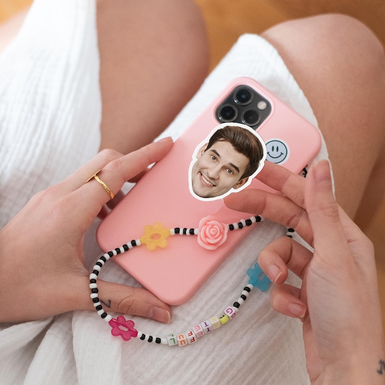 May include: A pink phone case with a sticker of a smiling face and a smiley face sticker. A beaded phone strap with flower and rose charms and the word "grateful" spelled out in letter beads. The phone is held in a person's lap.