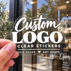 May include: A clear adhesive sticker featuring white text that reads Custom LOGO CLEAR STICKERS, with the additional text FULL COLOR, a heart icon, and ANY SHAPE. The sticker has a die-cut edge that follows the contour of the lettering and design.