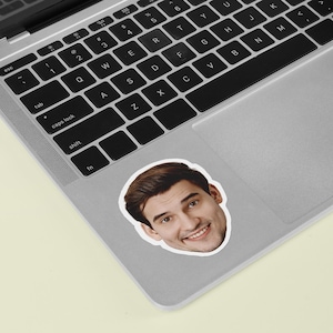 May include: A sticker of a smiling man's face, with brown hair and a white border. The sticker is on a silver laptop, with the keyboard visible. The background is a light yellow surface.