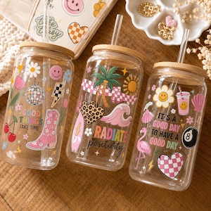 May include: Three clear glass tumblers with bamboo lids and glass straws, each featuring colorful decals. The first displays a disco ball and cowboy boot with the text "GOOD THINGS TAKE TIME." The second shows a palm tree and heart with "RADIATE positivity." The third features a flamingo and "IT'S A GOOD DAY TO HAVE A GOOD DAY."