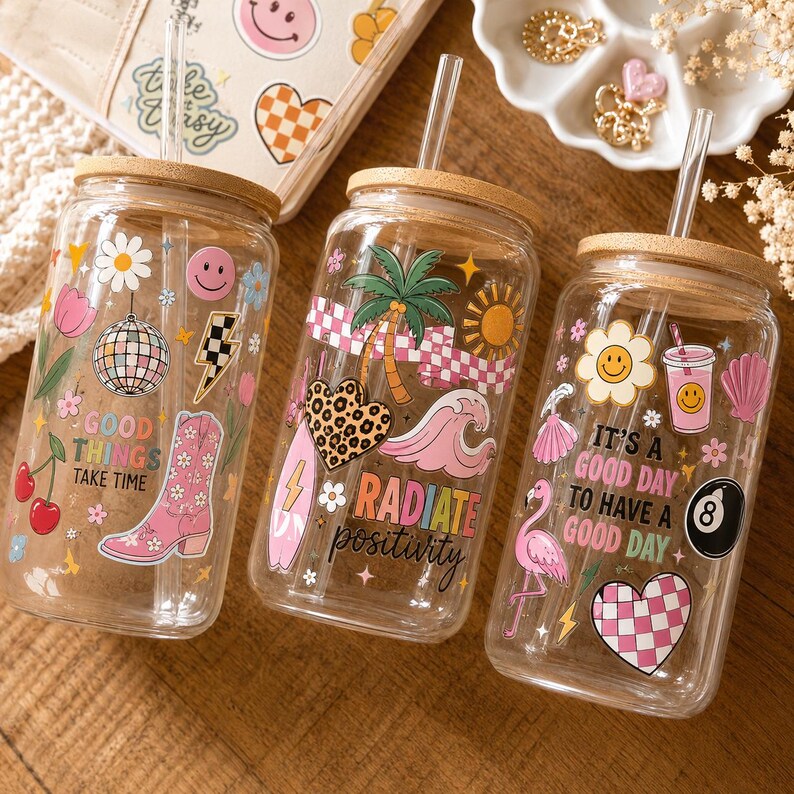 May include: Three clear glass tumblers with bamboo lids and glass straws, each featuring colorful decals. The first displays a disco ball and cowboy boot with the text "GOOD THINGS TAKE TIME." The second shows a palm tree and heart with "RADIATE positivity." The third features a flamingo and "IT'S A GOOD DAY TO HAVE A GOOD DAY."