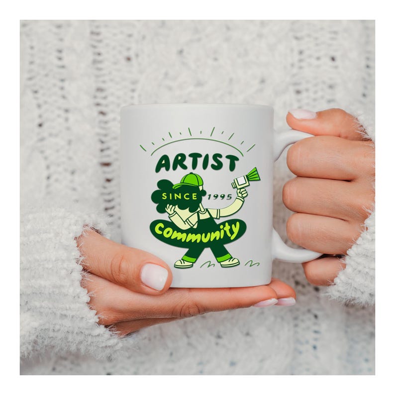 May include: White ceramic mug featuring a green and white cartoon design with the words "ARTIST COMMUNITY SINCE 1995". The design includes an artist character holding a paintbrush. The mug is held by a person wearing a white sweater.