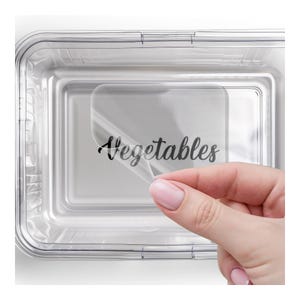May include: A clear plastic food storage container with a label that reads "Vegetables" in a handwritten font. The label is being peeled off, revealing the container's interior.