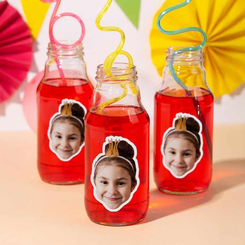 May include: Three glass bottles filled with red liquid, each with a colourful straw. Each bottle has a sticker of a smiling child wearing a crown. The background includes colourful paper decorations.