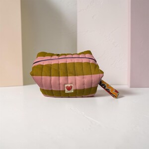 May include: A quilted zippered pouch featuring horizontal stripes in olive green and dusty pink. The pouch has a small square patch on the front with a red heart graphic and a patterned fabric pull tab attached to the side zipper closure.