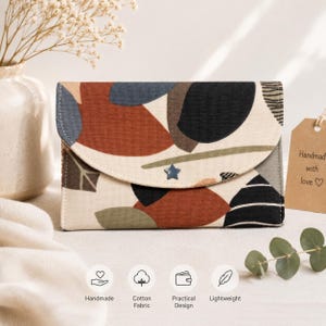 May include: A rectangular fabric wallet featuring a rounded flap closure with a small blue star-shaped stud. The wallet displays an abstract pattern of overlapping shapes in terracotta, navy blue, black, and olive green on a cream background.