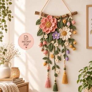 May include: A wall hanging featuring crocheted flowers and leaves suspended from a natural wooden branch. The arrangement includes a large pink flower, a white daisy, and smaller purple, yellow, and pink blossoms with green foliage and wooden beads.