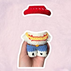 K&ouml;nnte beinhalten: Ein handgefertigtes H&auml;kel-Outfit, bestehend aus einem roten Cowboyhut und einem Miniatur-Shirt- und Shorts-Set. Das Shirt ist wei&szlig; mit gelben und roten Akzenten, und die Shorts sind blau mit einem braunen G&uuml;rtel. Das Outfit ist so gestaltet, dass es einem Charakter &auml;hnelt.