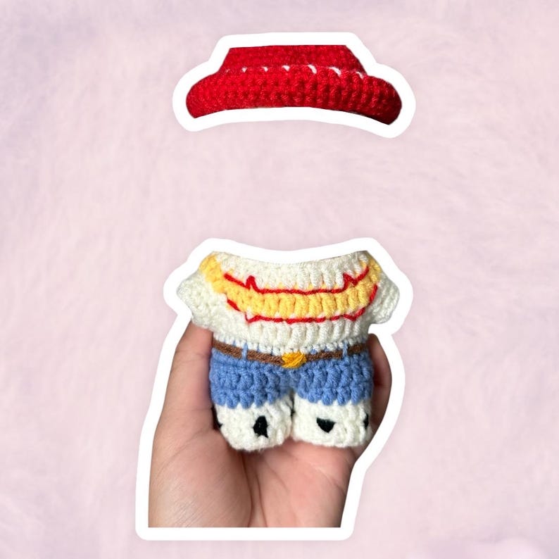 K&ouml;nnte beinhalten: Ein handgefertigtes H&auml;kel-Outfit, bestehend aus einem roten Cowboyhut und einem Miniatur-Shirt- und Shorts-Set. Das Shirt ist wei&szlig; mit gelben und roten Akzenten, und die Shorts sind blau mit einem braunen G&uuml;rtel. Das Outfit ist so gestaltet, dass es einem Charakter &auml;hnelt.