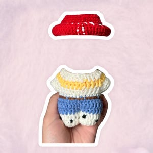 K&ouml;nnte beinhalten: Handgefertigtes H&auml;kel-Puppenoutfit in Rot, Wei&szlig;, Gelb und Blau. Das Set beinhaltet einen roten Hut und ein Oberteil mit gelben Streifen, kombiniert mit blauen Shorts. Die Puppenkleidung ist f&uuml;r ein kleines Spielzeug oder Sammlerst&uuml;ck.