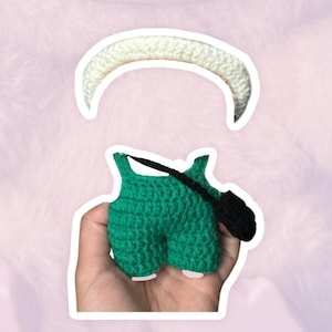 May include: A handmade crochet outfit in green, featuring shorts and suspenders, paired with a white crochet headband. The outfit is designed with a textured stitch, and the black suspenders add a contrasting detail.