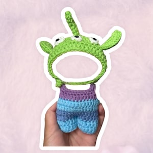 May include: A handmade crochet alien costume in green, purple, and blue. The costume features a green alien headpiece with three eyes and antennae, and a matching jumpsuit. The item is designed to be worn.