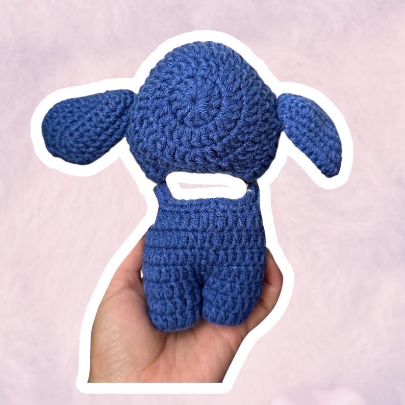 K&ouml;nnte beinhalten: Handgemachte blaue H&auml;kelpuppe mit gro&szlig;en Ohren und einem Overall. Die Puppe wird in einer Hand vor einem weichen rosa Hintergrund gehalten. Die H&auml;kelstiche sind sichtbar und verleihen der Puppe ein strukturiertes Aussehen.