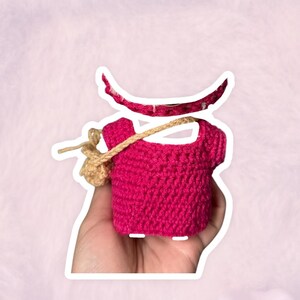 Puede incluir: Un top de ganchillo rosa intenso hecho a mano con una peque&ntilde;a bolsa de ganchillo beige sujeta con una correa beige. Una forma de media luna rosa intenso a juego est&aacute; encima del top. El trabajo de ganchillo es visible y detallado.
