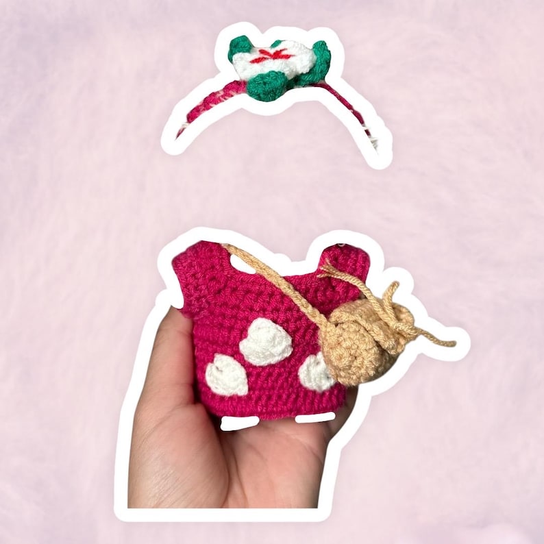 Puede incluir: Un conjunto de ganchillo hecho a mano con una diadema con una flor verde y blanca y una parte superior magenta con lunares blancos. La parte superior tiene un accesorio en forma de hongo de color beige.