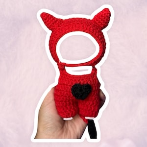 May include: A handmade red crochet outfit with a devil horn hood and a black heart on the back. The outfit is designed to be worn, with openings for the face and legs. The item is held in a hand.