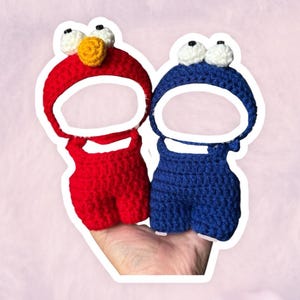 May include: Two crocheted character-themed baby outfits. One is red with a yellow nose and white eyes, the other is blue with white eyes. Both have open face areas and are designed to be worn by a baby.