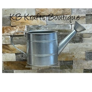 May include: A galvanized metal watering can with a handle and spout. The watering can is cylindrical with a silver finish. The text "KB Krafts Boutique" is above the watering can. The background is a stone wall.