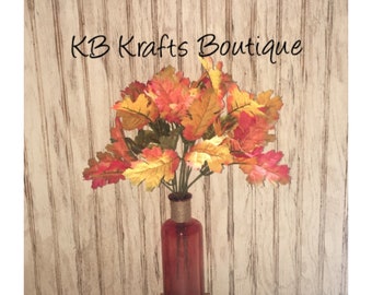 May include: A bouquet of autumn-colored leaves in a red glass bottle. The leaves display shades of red, orange, and yellow. The bottle is wrapped with twine at the neck. The text "KB Krafts Boutique" is visible.